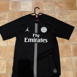 Svart PSG fotbollströja Jordan - Snygg svart PSG fotbollströja från Jordan-kollektionen med Fly Emirates-tryck, PSG-logga och Jordan-logga på bröstet. Vertikal grå rand framtill, korta ärmar och namn/nummer (JAMIE 10) på ryggen. Tillverkad i lätt och ventilerande material.