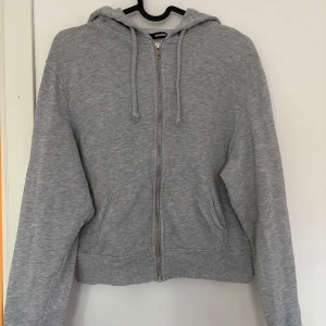 Grå zip hoodie  - Säljer en grå zip hoodie från Bikbok med dragkedja framtill, huva med snörning och två fickor. Tröjan är lite croppad och har långa ärmar. 