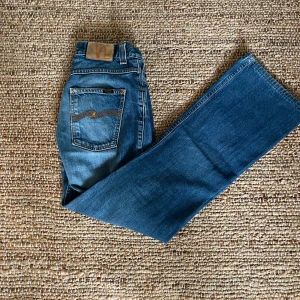 Blå bootcut jeans från Nudie - Snygga blå jeans från Nudie med klassisk bootcut-passform och coola detaljer på bakfickan. Jeansen har en mellanblå tvätt och är tillverkade i slitstarkt denim. Perfekta för dig som gillar en tidlös och avslappnad stil.