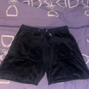 Svarta velourshorts Juicy Couture - Svarta shorts i mjuk velour från Juicy Couture med snörning i midjan och stora fickor både fram och bak. Klassisk broderad JC-logga på ena fickan och detaljer i silver. Perfekta för en chill och trendig look.