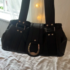 Svart axelväska med hästsko-detalj - Cool svart axelväska i skinnimitation med breda axelband och silvriga nitar. Väskan har en stor metallhästsko framtill och dragkedja upptill. Perfekt för dig som gillar unika detaljer och vill sticka ut lite extra. Den rymmer jätte mycket.💓😊