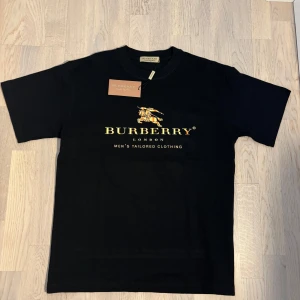 Svart t shirt - Svart t-shirt från Burberry med klassisk guldfärgad logga och text på bröstet. Rak passform och rund halsringning. Tillverkad i mjuk bomull, perfekt för en clean och stilren look. Storlek ”s”