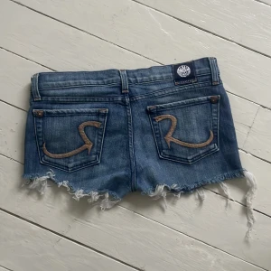 Rock & Republic jeansshorts med rå kant - Snygga blå jeansshorts från Rock & Republic med slitna, fransiga kanter och klassisk femficksdesign. Bakfickorna har broderade R-detaljer. Perfekta för en avslappnad look under varma dagar. Storlek 28 och stretchar sig 