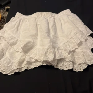 Vit volangkjol från H&M Divided - Supersöt vit kortkjol från H&M Divided med broderade volanger och elastisk midja. Kjolen har romantiska spetsdetaljer och är perfekt för en somrig look. Tillverkad i bomull för en luftig och bekväm känsla.