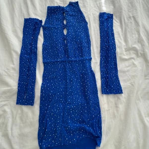 Blå glittrig kortklänning med ärmar - Snygg blå kortklänning med glittriga prickar och avtagbara ärmar. Klänningen har snörningsdetaljer framtill och är figurnära. Perfekt för dig som vill sticka ut med en unik look.