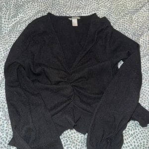 Svart omlottopp från H&M - Svart omlottopp från H&M med lång ärm och snygg rynkdetalj framtill. Toppen har v-ringning och är tillverkad i ett mjukt, stretchigt material som sitter skönt på kroppen. Perfekt att styla med jeans eller kjol för en trendig look.