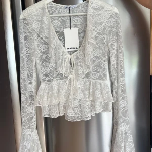 Vit spetsblus med volang från Bershka - En jättefin vit blus i spets från Bershka med långa trumpetärmar, volangdetaljer och knyt framtill. Blusen är genombruten med blommigt spetsmönster och har v-ringning. Blusen är helt oanvänd och kostade 30€ i Malaga. 