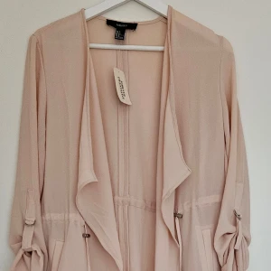 Beige/ljusrosa oversize trenchcoat med snörning - Snygg beige/ljusrosa trenchcoat i oversized modell med dragsko i midjan och vid fållen. Jackan har långa ärmar som kan rullas upp och fästas med knapp, stora fickor framtill. Öppen modell utan knäppning. Helt ny – med originaltag kvar. Storlek S