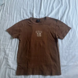 Brun Baby t-shirt - Urban Outfitters - Brun t-shirt från Urban Outfitters med trycket 'Colorado Springs' i vitt framtill. Klassisk rund hals och korta ärmar. Skön och enkel modell i bomull, perfekt till jeans eller shorts.