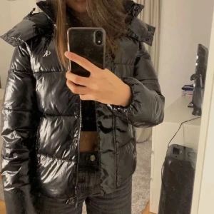 Svart pufferjacka från Calvin Klein - Snygg svart pufferjacka från Calvin Klein med glansig finish och diskret CK-logo på bröstet. Jackan har hög krage, huva och dragkedja framtill. Perfekt för kalla dagar och ger en cool vibe till din outfit.