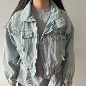 Ljusblå jeansjacka med bälte - Snygg ljusblå jeansjacka med dragkedja, stora bröstfickor och bälte i midjan. Jackan har en loose passform, elastiska ärmslut och coola detaljer som snörning vid kragen. Perfekt för dig som gillar en avslappnad och trendig stil.