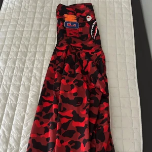 BAPE röd camo durag med shark-tryck - Snygg durag från BAPE i röd och svart camo med ikoniskt haj- och tigertryck samt broderade bokstäver. Tillverkad i glansig polyester som ger en clean look. Perfekt accessoar för att lyfta din streetwear-stil.