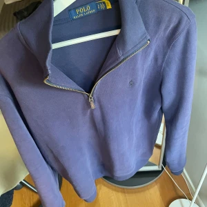 Lila half zip tröja från Polo Ralph Lauren - Snygg lila långärmad tröja från Polo Ralph Lauren med half zip-dragkedja och klassisk krage. Tröjan är i bomull och har en broderad logga på bröstet. Perfekt för dig som gillar stilrena och bekväma plagg.