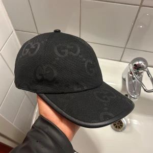 Svart keps från Gucci med GG-mönster - Snygg svart keps från Gucci med klassiskt GG-monogram i ton-i-ton. Kepsen har böjd skärm, justerbar rem bak och är tillverkad i slitstark bomullscanvas med detaljer i läder. Perfekt för dig som vill ha en stilren och lyxig accessoar.
