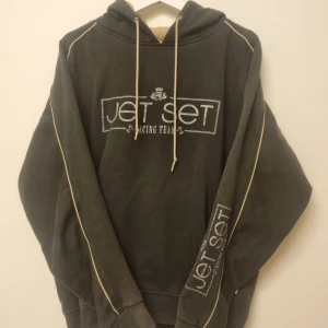 Vintage hoodie 90-tal - Mörkgrön hoodie från Jet Set Racing Team med vita kontrastsömmar längs ärmarna och tryck på bröstet och ena ärmen. Tröjan har huva med snörning och ribbade muddar. Perfekt för dig som gillar streetwear och sportig stil.