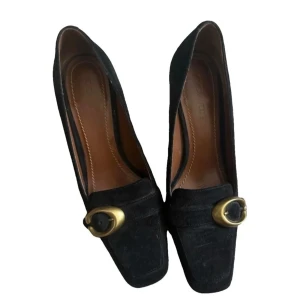 Svarta mocka pumps från Coach - Stilrena svarta pumps i mocka från Coach med fyrkantig tå och chunky klack. Snygg detalj med guldfärgat spänne framtill. Insida i brunt skinn för extra komfort. Perfekta för dig som vill ha en klassisk men trendig look.