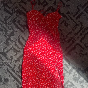 Röd blommig sommarklänning med slits - Supersöt röd klänning med vita småblommor, smala axelband och snörning framtill. Klänningen är figurnära och har en slits på sidan som ger en extra touch. Perfekt för varma dagar och har en romantisk vibe.