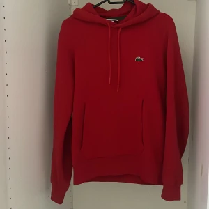 Lacoste hoodie - Snygg röd hoodie från Lacoste i klassisk passform. Tröjan har huva med snörning, stor magficka och det ikoniska krokodilmärket på bröstet. Perfekt för dig som gillar stilrena och sportiga vibes.