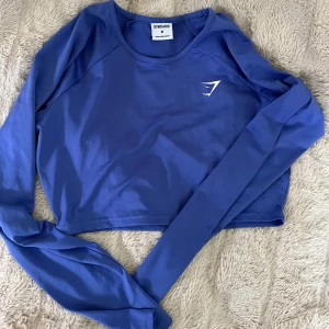 Blå croppad långärmad tröja Gymshark - Säljer en blå croppad långärmad tröja från Gymshark. Stretchig och sval polyestermaterial. Tröjan har en stilren design med Gymshark-logga på bröstet och rund halsringning. Superskön. 
