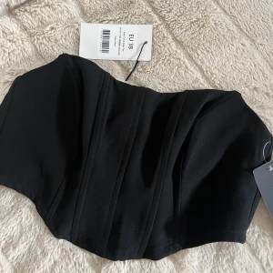 Svart korsettopp från NA-KD - Säljer en svart korsettopp från NA-KD med markerade sömmar och snygg bandeauform. Toppen är axelbandslös och har en stilren, figurnära passform som ger en cool och trendig look. Perfekt att styla med höga jeans eller kjol. aldrig använd. 