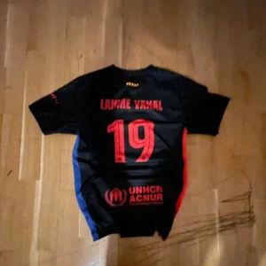 FC Barcelona Lamine Yamal tröja - Snygg svart fotbollströja från FC Barcelona med Lamine Yamal och nummer 19 på ryggen. Tröjan har röda detaljer, blå sidopanel och klubbmärke på bröstet. UNHCR-logga tryckt nedtill. Tillverkad i lätt och ventilerande material, perfekt för match eller träning.