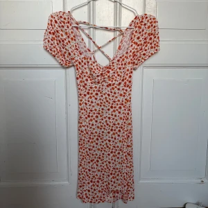 Vit blommig långklänning med snörning - Säljer en superfin långklänning med vit bas och röda blommor. Klänningen har puffiga korta ärmar, sweetheart liknande neckline och snygg snörning i ryggen som ger en cool detalj. Materialet känns lätt och flowy, perfekt för varma dagar. Passformen är normal/tajt och klänningen har en feminin siluett. Klänningen e väldigt stretchig så kan passa många!