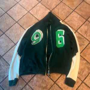 Grön stickad bomberjacka J.Lindeberg - Säljer en grön stickad bomberjacka från J.Lindeberg med vita ärmar och coola broderade siffror på framsidan. Jackan har ribbade muddar, dragkedja och en schysst loose passform. Perfekt för dig som vill sticka ut med sportig stil och snygga detaljer.