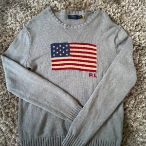 Grå stickad tröja Polo Ralph Lauren - Säljer en grå stickad tröja från Polo Ralph Lauren med amerikansk flagga i blått, rött och vitt på bröstet samt RL-broderi. Tröjan har rund hals och långa ärmar med ribbade muddar. Det står S på tröjan men sitter som M. Perfekt för dig som gillar klassisk och cool stil men även en avslappnad look.Har du några funderingar så är det bara att skriva👌