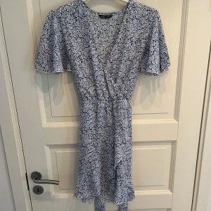 Fin blommig omlottklänning  - Säljer en superfin ljusblå omlottklänning från SHEIN med vita blommor, korta ärmar och volangkant nedtill💕💕Klänningen har justerbart knytband i midjan och v-ringad hals. Jätte fin passform och knappt använd!!🫶🏼🫶🏼