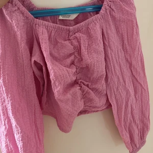 Rosa croppad blus från H&M - Superfin ljusrosa croppad blus från H&M med långärmade puffärmar och resår vid axlar och ärmslut. Blusen har en rynkad detalj framtill och är gjord i ett krinklat, lätt material som känns luftigt och skönt. Perfekt för dig som gillar trendiga och bekväma plagg.