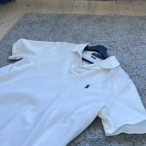 Vit pikétröja från Polo Ralph Lauren - Klassisk vit pikétröja från Polo Ralph Lauren med blå broderad logga på bröstet. Tröjan har korta ärmar, krage och knappslå. Classic fit-modell i bomull som är både stilren och enkel att matcha.