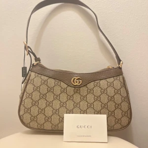 Gucci axelväska i beige canvas - Säljer en snygg axelväska från Gucci i beige canvas med klassiskt GG-mönster och bruna skinn-detaljer. Väskan har guldfärgad GG-logga framtill och dragkedja upptill. Perfekt storlek för det viktigaste och en ikonisk accessoar.