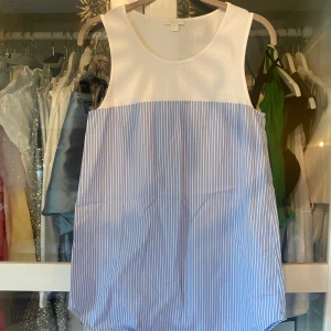 Linne /topp/kortärmad blus COS - Säljer en ärmlös topp eller kortklänning från COS i storlek 38. Överdelen är vit och ribbad, medan nederdelen är ljusblå med vita ränder. Klänningen är tillverkad i 100% bomull och har en loose passform som ger en chill vibe. Perfekt för varma dagar och enkel att styla med sneakers eller sandaler.