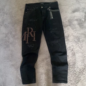 Svarta jeans från Amiri med patch - Säljer ett par svarta jeans från Amiri med bruna, stora bokstavspatchar på både fram- och baksida där det står AMIRI. Jeansen har distressed detaljer och en slim passform. Är nya