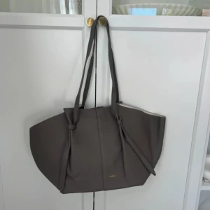 Grå Väska  i skinn - Snygg och rymlig väska i grått skinn med stilrena knyt-detaljer på handtagen. Väskan har en mjuk, strukturerad form och insidan är ljusbeige med magnetknäppning. Perfekt för dig som gillar minimalistisk design och vill ha plats för allt.