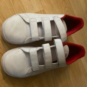 Vita adidas sneakers med kardborre - Säljer ett par vita adidas sneakers med röd insida och tre smidiga kardborreband. Skorna har en klassisk rund tå och platt sula, med adidas-logga i rött på plösen. Perfekta för dig som gillar enkel och sportig stil.
