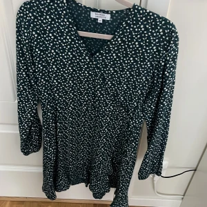 Prickig grön kortklänning Galeries Lafayette - Snygg kortklänning från Galeries Lafayette i mörkgrönt med vita prickar. Klänningen har v-ringad hals, långärmade ärmar och en lätt volang vid nederkanten. Perfekt för dig som gillar mönstrade plagg med lite extra attityd.