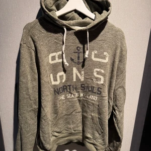 Grön hoodie från North Sails - Snygg grön hoodie från North Sails med vit snörning i huvan och tryck framtill. Tröjan har känguruficka och långa ärmar, perfekt för en avslappnad stil. Mjuk bomullsblandning som känns skön mot huden.