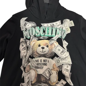 Svart Moschino hoodie med nalletryck - Svart hoodie från Love Moschino med stort tryck av en nalle, dollar bills och texten 'This is not a Moschino toy' på framsidan. Tröjan har huva och långa ärmar, perfekt för dig som gillar statement-plagg och streetstyle.