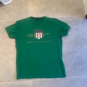 Grön GANT t-shirt med tryck - Snygg grön t-shirt från GANT med stort tryck på bröstet och texten 'American Sportswear'. Klassisk rund hals och korta ärmar. Perfekt för dig som gillar sportig och stilren look. Gjord i mjuk bomull för skön känsla hela dagen.
