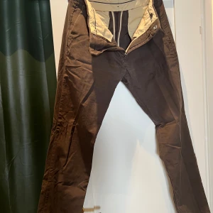 Bruna chinos från NN07, stl 32/32 - Snygga bruna chinos från NN07 i modellen Theo 1500. Byxorna har raka ben, klassiska fickor och är tillverkade i mjuk bomull med lite stretch för extra komfort. Perfekta för en clean och stilren look.