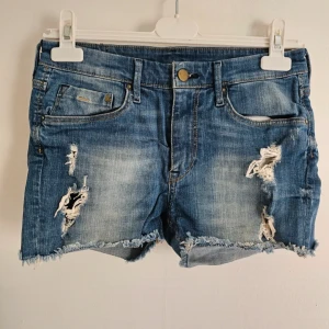 Blå jeansshorts med slitningar H&M - Snygga blå jeansshorts från H&M med rå kant och slitna detaljer framtill. Klassisk femficksmodell med knapp och dragkedja. Materialet är 98% bomull och 2% elastan för skön stretch. Perfekta för sommarens chill dagar.