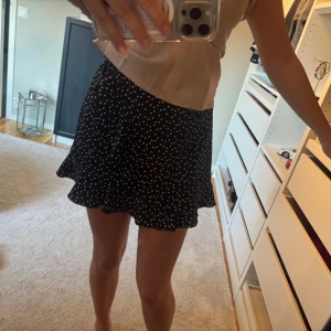 Svart kortkjol med vita hjärtan - Supersöt svart kortkjol med vita hjärtan som mönster. Kjolen har en lätt volang nedtill och man knyter kjolen med en roset vid sidan. 