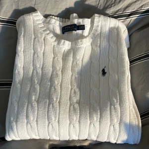 Vit kabelstickad tröja Polo Ralph Lauren - Vit kabelstickad tröja från Polo Ralph Lauren med klassisk rund halsringning och broderad marinblå logga på bröstet. Tröjan är i mjuk bomull och har långa ärmar. Perfekt för dig som gillar en clean och tidlös stil.