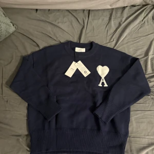Mörkblå stickad tröja från Ami - Snygg mörkblå stickad tröja från Ami Alexandre Mattiussi med vit hjärtlogga på bröstet. Tröjan har rund hals, långa ärmar och breda ribbade muddar vid ärmslut och nederkant. Perfekt för dig som gillar stilrena och trendiga plagg.
