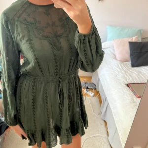 Mörkgrön kort klänning med volang - Supersöt mörkgrön kort klänning med långa ärmar, volangkant nertill och broderade detaljer. Klänningen har knyt i midjan och transparent mesh upptill med broderat mönster. Perfekt för dig som gillar boho-vibes och vill ha något unikt.