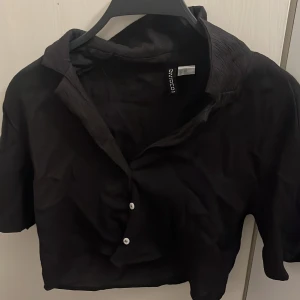Svart croppad skjorta från H&M Divided - Svart croppad skjorta från H&M Divided med korta ärmar och två vita knappar framtill. Skjortan har en klassisk krage och är tillverkad i ett lätt, lite skrynkligt tyg som ger en avslappnad vibe. Perfekt för dig som gillar en stilren och enkel look.