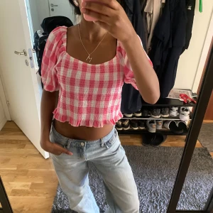 Rutig rosa croppad blus med puffärm - Superfin croppad blus i rosa och vitt med rutigt mönster och puffiga korta ärmar. Toppen har smockad struktur, volangkant nertill och kan bäras både på axlarna eller offshoulder. Perfekt för en söt och trendig sommarlook! 
