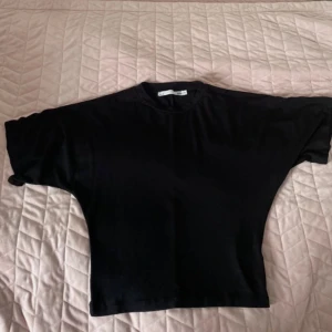 Svart croppad t-shirt - En svart croppad t-shirt med korta ärmar och rund hals. T-shirten har en loose passform och är tillverkad i mjuk bomull, perfekt för en chill och avslappnad stil. Enkel design utan mönster eller detaljer, passar till jeans eller shorts.