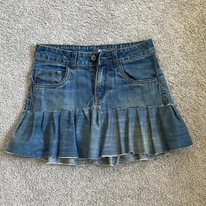 Blå jeanskjol med volang - Säljer en blå jeanskjol med volangkant och klassiska fickor både fram och bak. Kjolen har bälteshällor, knapp och dragkedja framtill. Den är kort och har en cool, avslappnad vibe med råa kanter på volangen. Perfekt för dig som gillar denim och vill sticka ut.